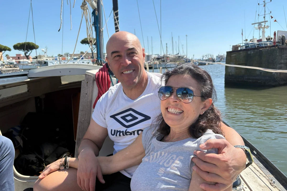 Antonio and Rita smiling on board a moored sailing boat, with the ropes and the other boats of the harbour in the background.